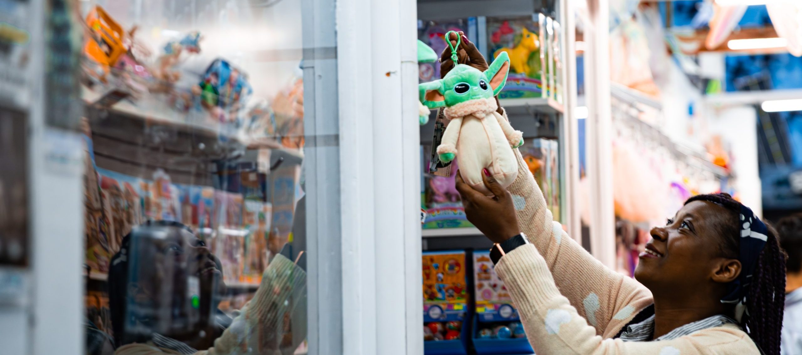 A woman smiles as she reaches for a Baby Yoda plush on a high shelf in a toy store.