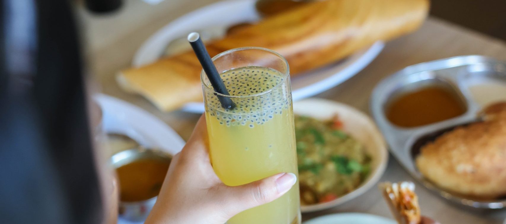 A person holds a yellow drink with seeds on top while enjoying South Indian vegetarian dishes at a restaurant.